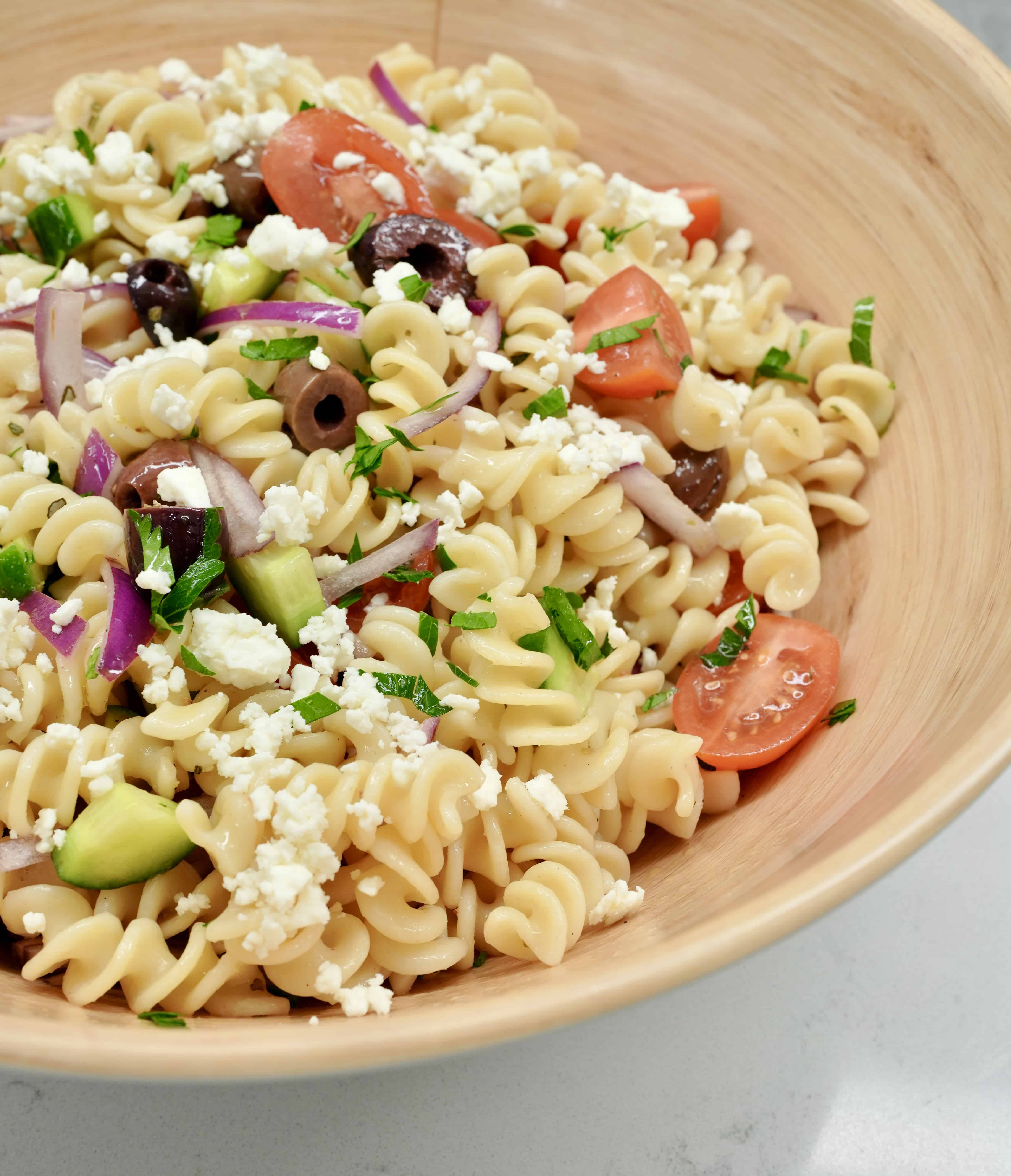 Large serving bowl filled with Greek pasta salad: rotini pasta, sliced tomatoes, olives, red onions, and parsley