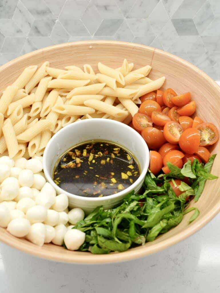 Large bowl made up of ingredients for pasta salad: penne pasta, sliced cherry tomatoes, chopped basil, mozzarella balls, and olive oil balsamic glaze.