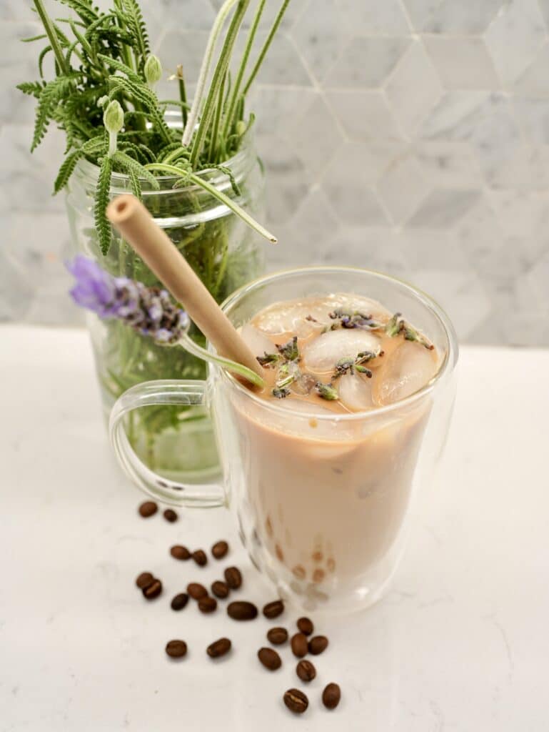 Iced lavender latte served in glass mug, next to jarred lavender plant and coffee beans