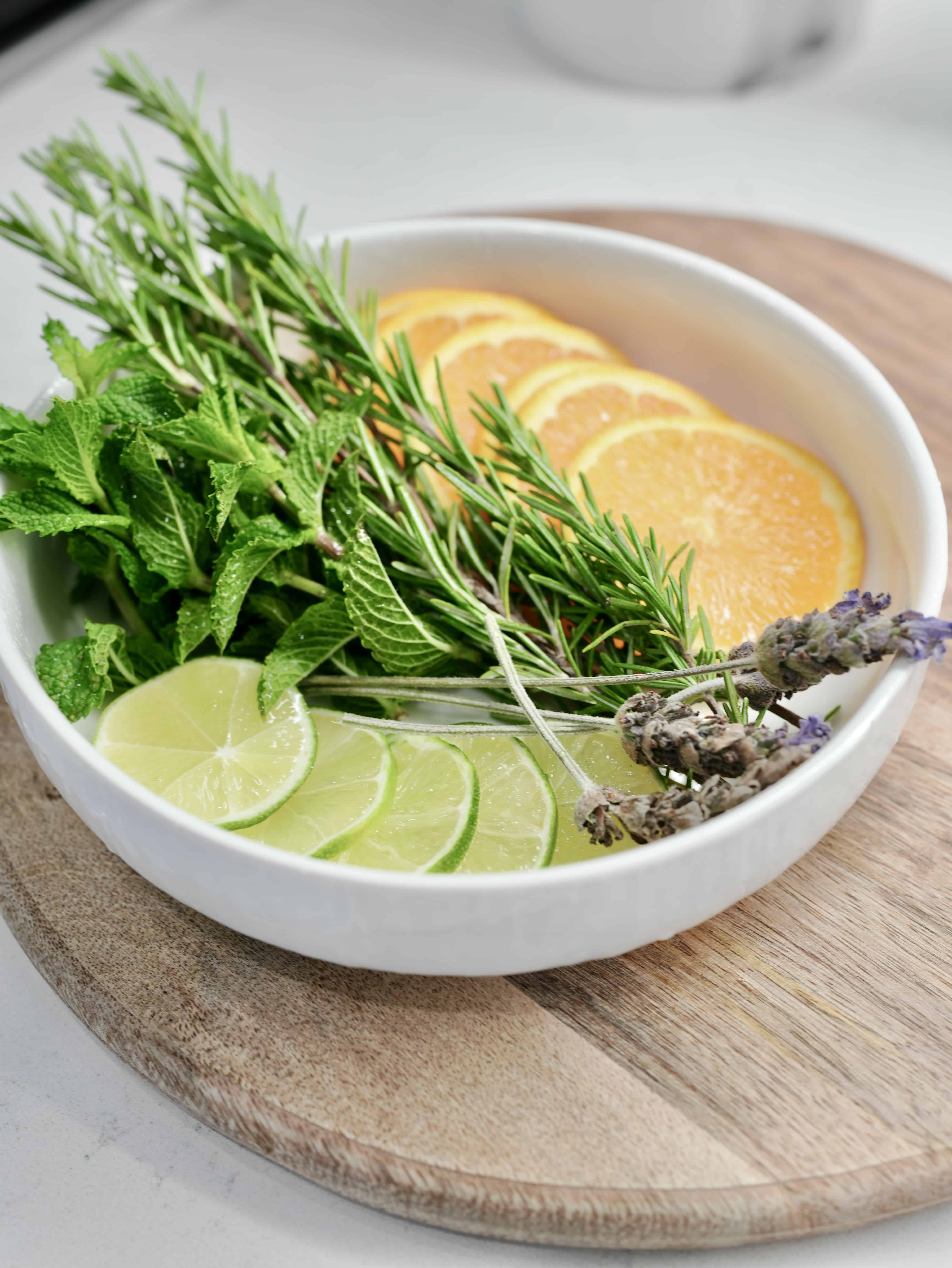 White bowl with orange and lime slices, lavender flowers, rosemary sprigs, and mint on wooden board