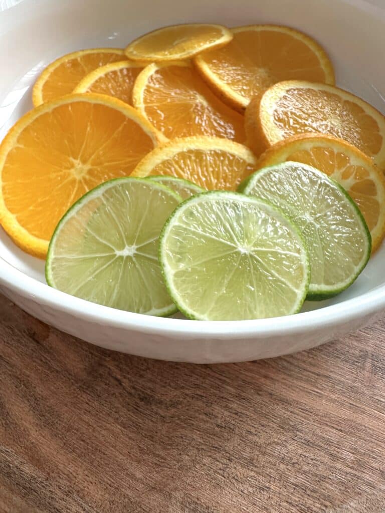 Fresh slices of orange and lime in white bowl on wooden cutting board