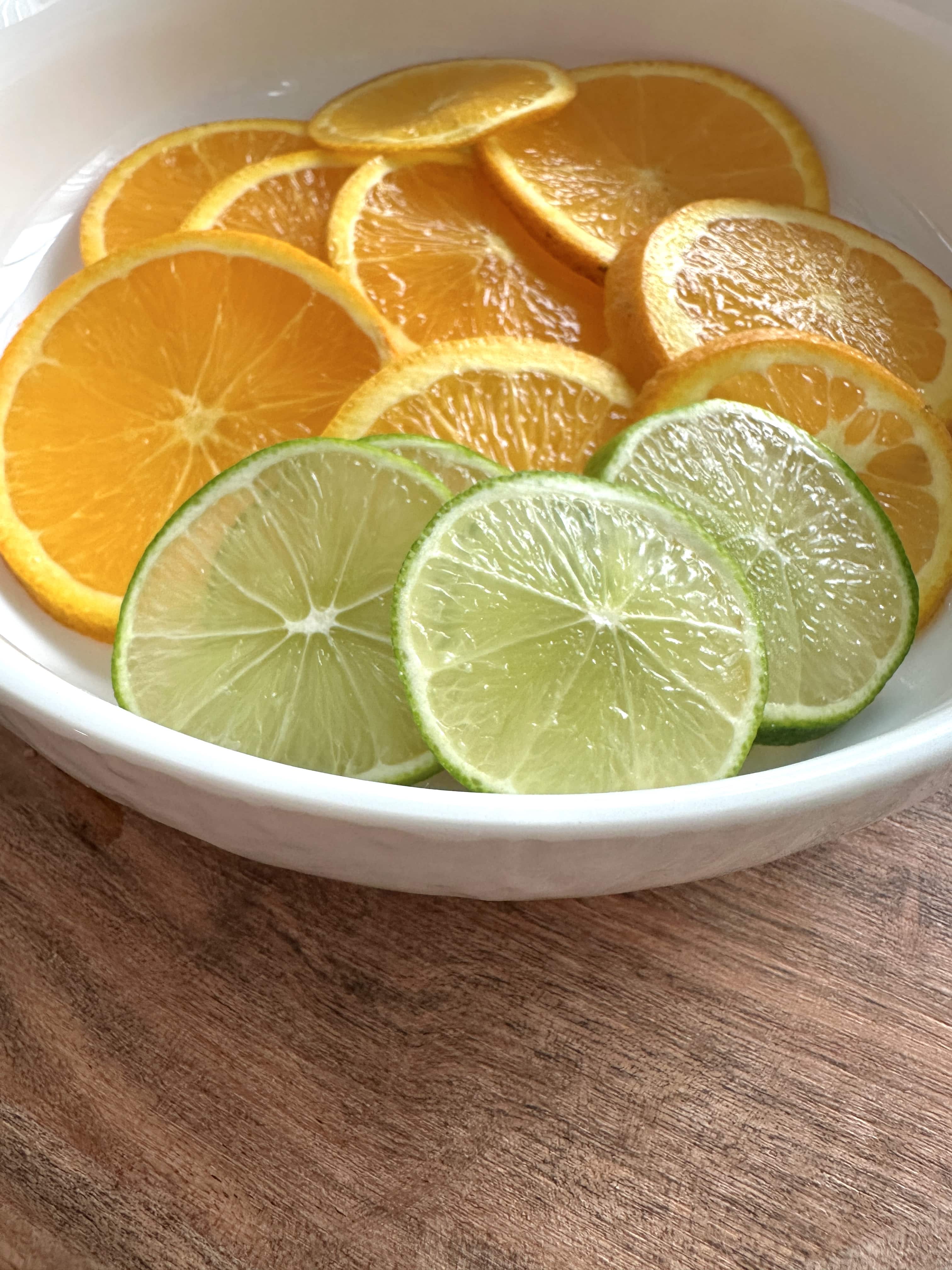 Fresh slices of orange and lime in white bowl on wooden cutting board
